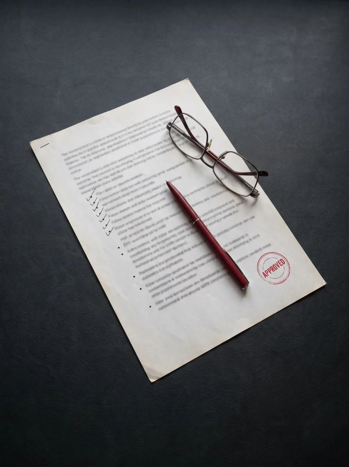 Bedrucktes Dokument mit rotem APPROVED-Stempel, dunkelrotem Stift und Lesebrille darauf.